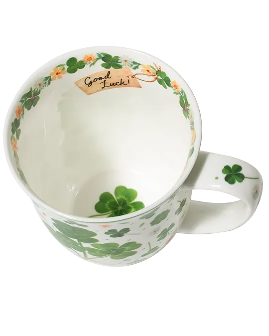 White mug with green four-leaf clovers tied in a bouquet, small flowers, and a pale green rim.