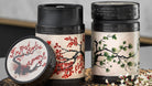Two Akashi tea tins with black lids and cream backgrounds, one featuring red blossoms and the other green pine branches.