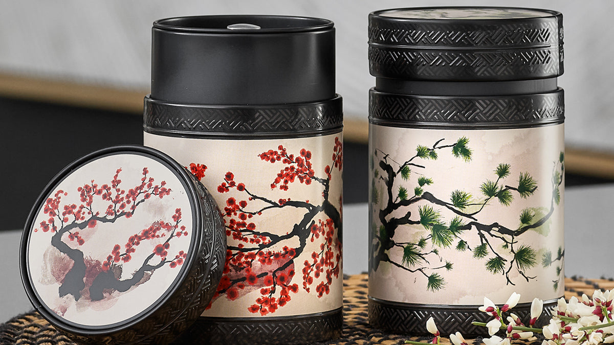 Two Akashi tea tins with black lids and cream backgrounds, one featuring red blossoms and the other green pine branches.